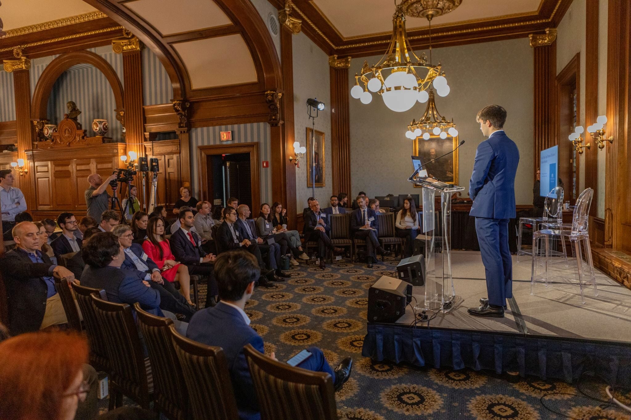 A person in a suit is speaking at a podium with a laptop in an ornate room, addressing an audience seated in wooden chairs. The setting includes chandeliers and decorative wood paneling.