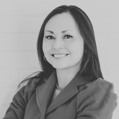 A grayscale headshot of a smiling person with long hair, wearing a collared jacket and a necklace.