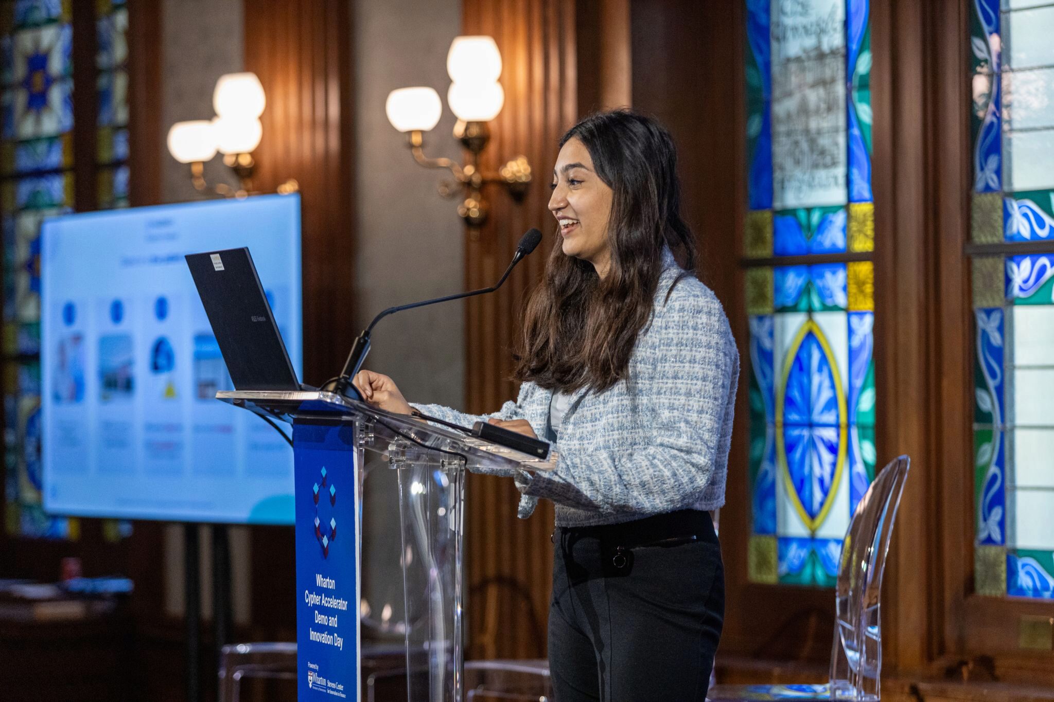 A person is speaking at a podium with a laptop, in front of a screen displaying presentation slides. The setting includes stained glass windows and elegant lighting, indicating a formal event or conference.